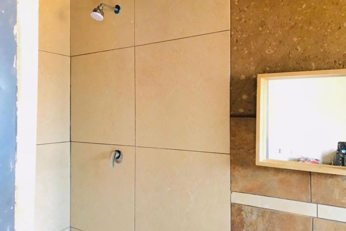 A bathroom area featuring large format beige wall tiles, a shower head, and a wooden-framed mirror, currently in an unfinished state.