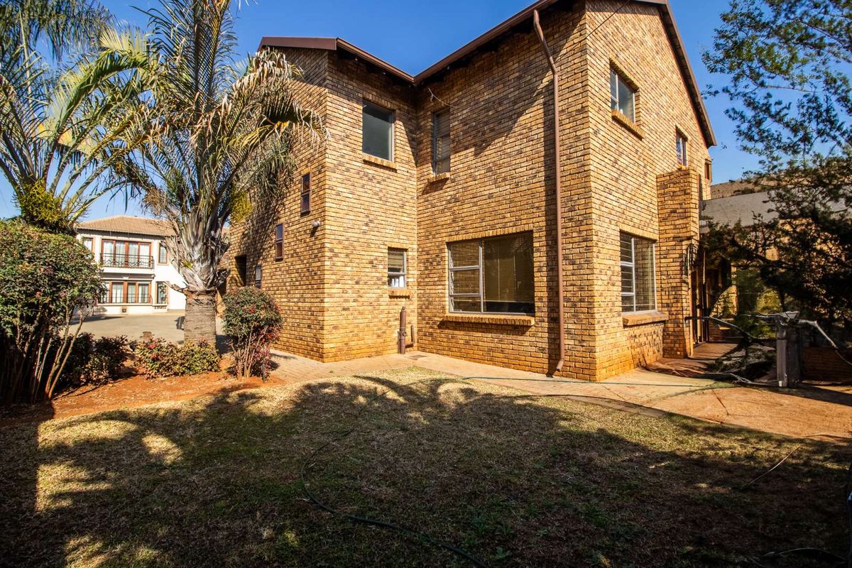 A view of the rear exterior of a multi-story brick house featuring established landscaping and a paved walkway.