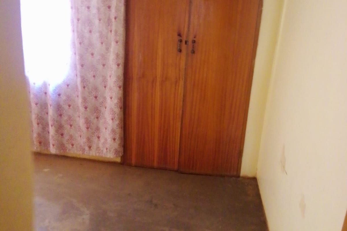 A bedroom featuring built-in wooden cupboards and a window with curtains, requiring cosmetic updates to the flooring and wall finishes.