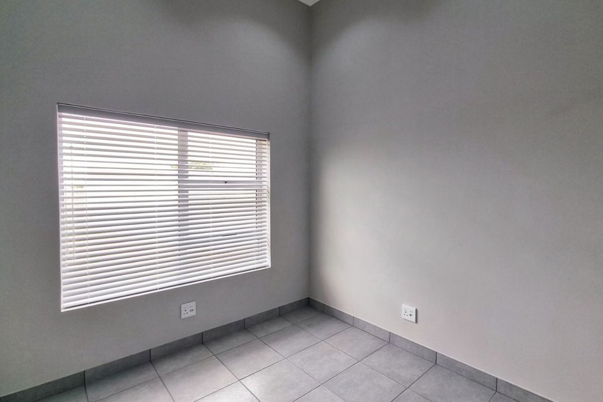A clean, empty room featuring neutral grey floor tiles, plain walls, and a window fitted with horizontal blinds.