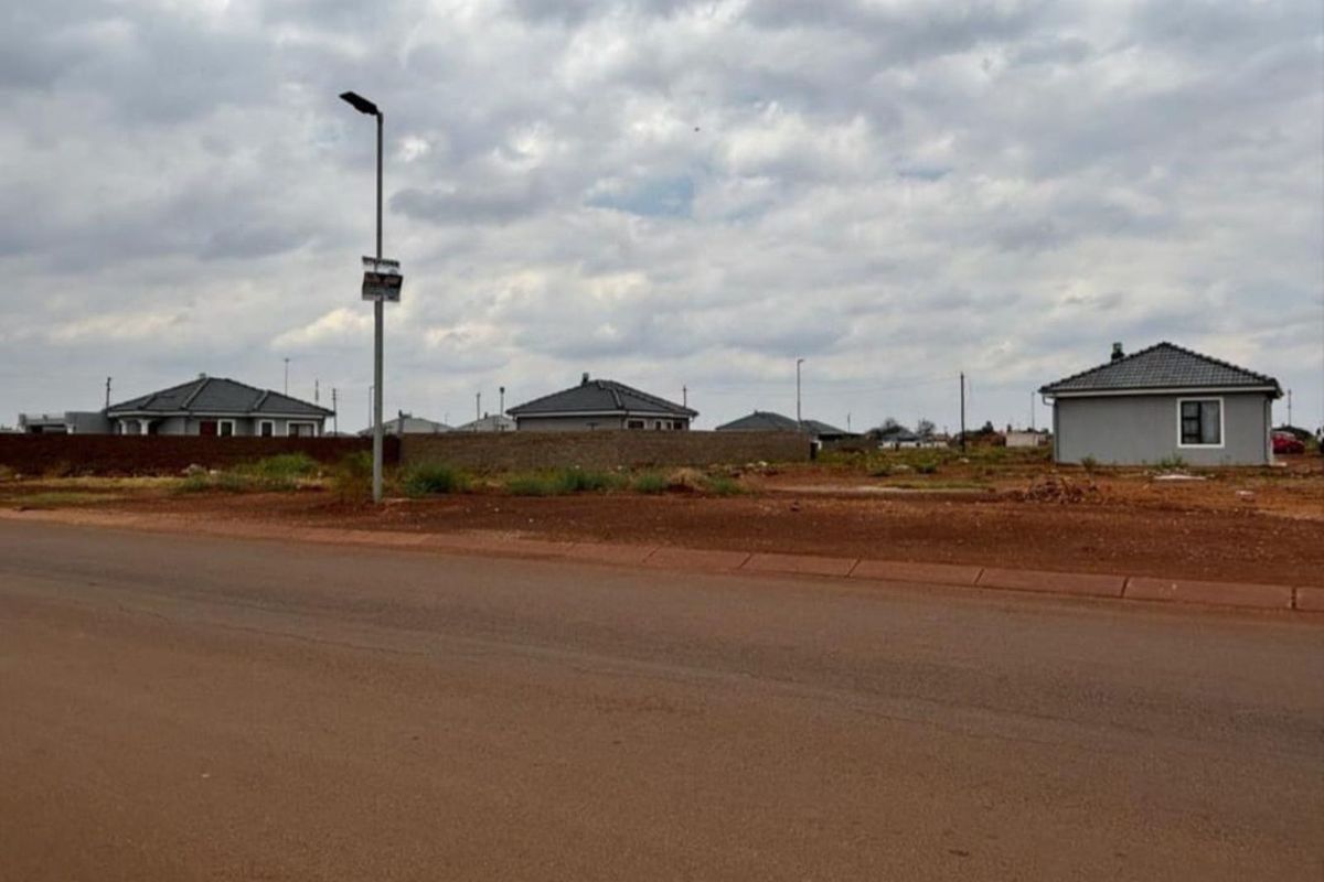 A street-level view of a vacant residential plot featuring red soil, adjacent to established neighborhood housing and a street lamp.