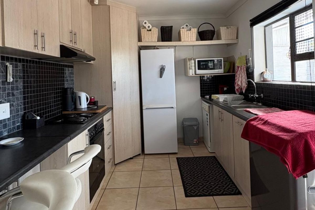 A functional galley-style kitchen featuring light wood-look cabinetry, black tiled splashbacks, dark countertops, and tiled flooring with integrated appliance spaces.