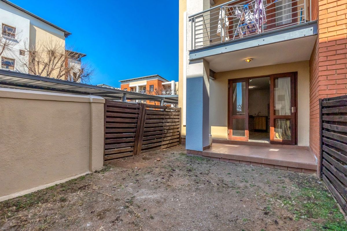 A ground floor apartment patio area featuring tiled flooring, a small private garden space, and a balcony above with metal railings.