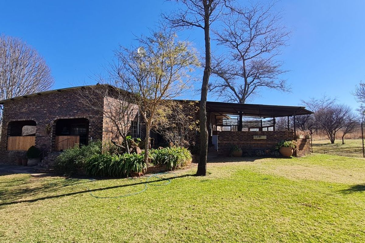 A rustic face-brick farmhouse exterior featuring a covered patio area, set within a spacious, established garden landscape.