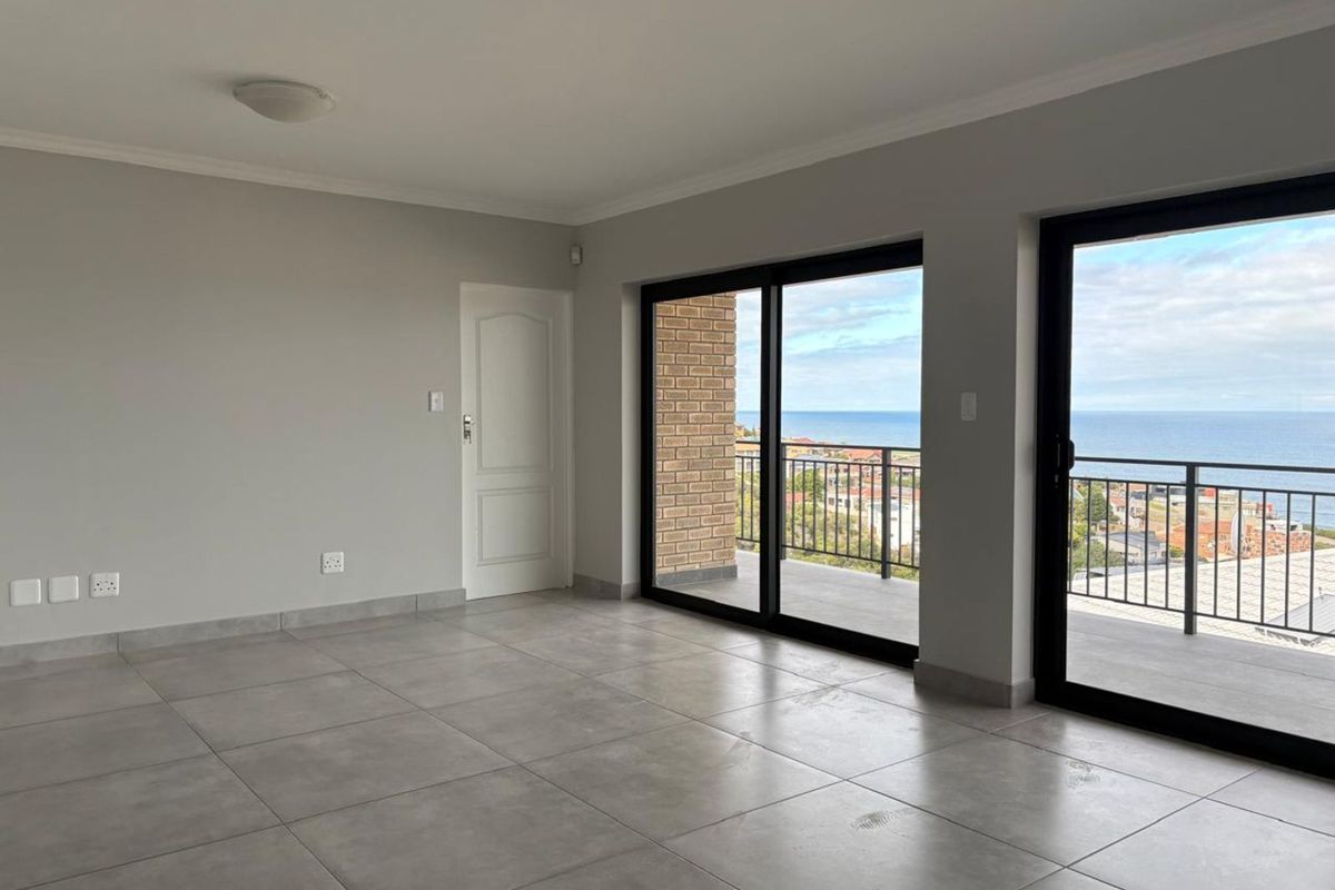 A bright, empty living area featuring large sliding glass doors that open onto a balcony with expansive ocean views and tiled flooring throughout.