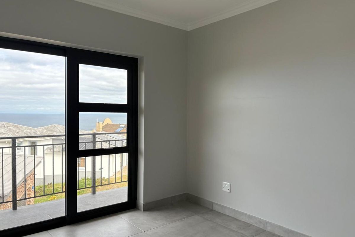 A bright, empty bedroom featuring large sliding glass doors that open onto a balcony with coastal views and modern tiled flooring.