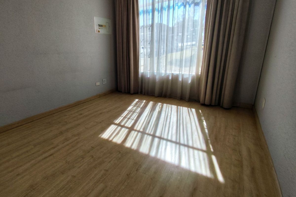 A bright, empty bedroom featuring light wood-look flooring, neutral walls, and large windows dressed with floor-to-ceiling curtains.
