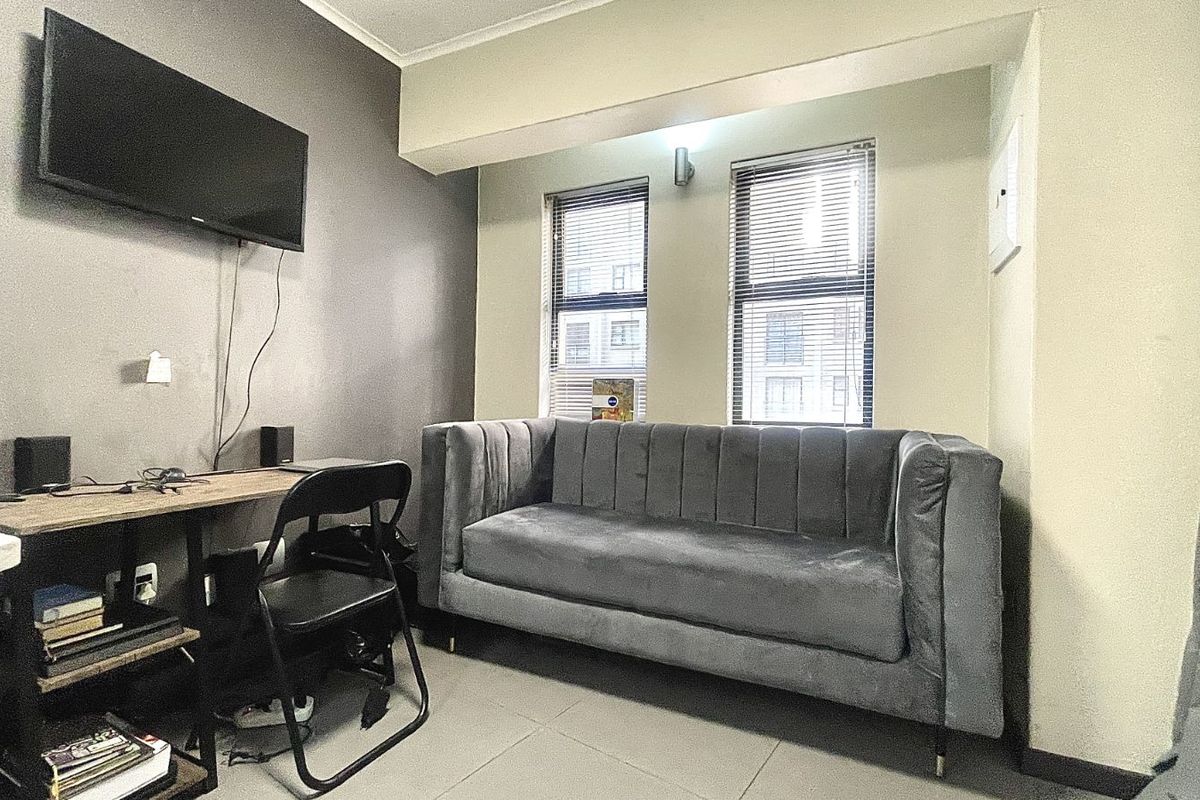 A compact living room featuring tiled flooring, neutral wall tones, and dual windows with fitted blinds, providing a functional and well-lit space.