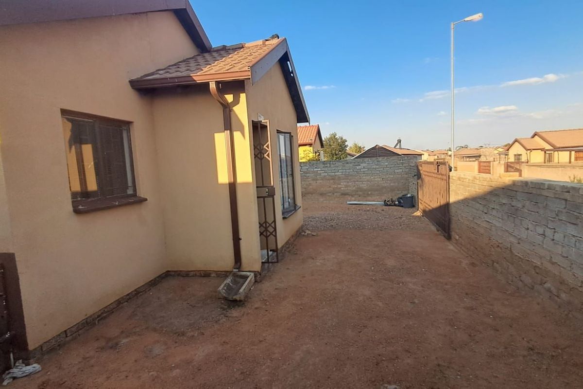 A view of the rear exterior of the house, featuring a simple facade, tiled roof, and an unpaved backyard area enclosed by boundary walls.