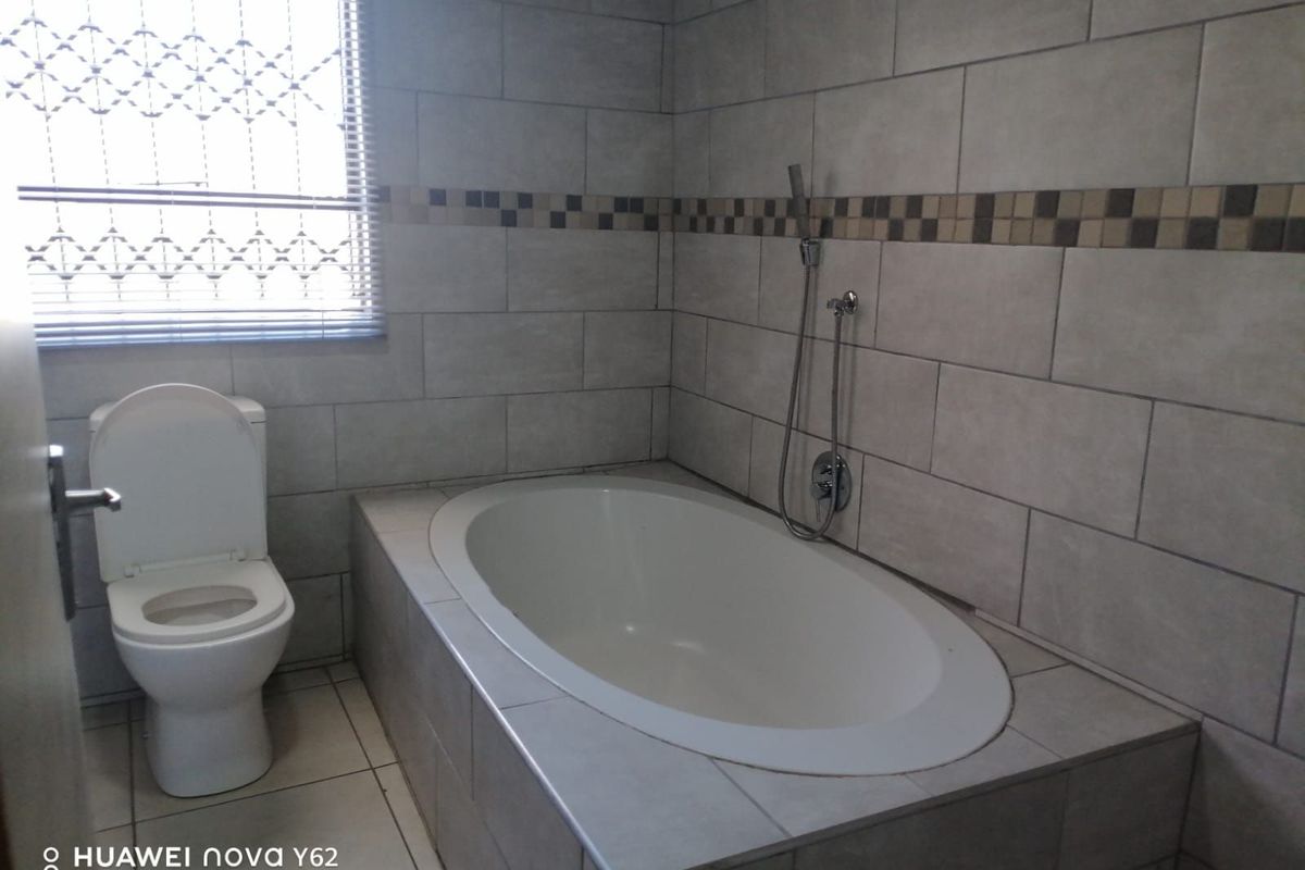 A functional bathroom featuring a built-in oval bathtub with a handheld shower mixer and a standard toilet, finished with neutral wall and floor tiling.