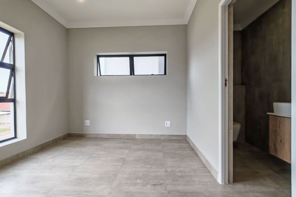 A bright, empty bedroom featuring neutral tiled flooring, modern recessed lighting, and an en-suite bathroom with a floating vanity.