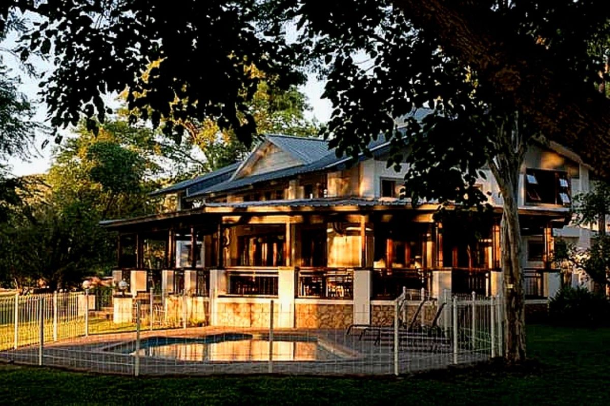 A scenic view of the farmhouse exterior featuring a wrap-around veranda, stone-based pillars, and a fenced swimming pool area set within a lush garden.