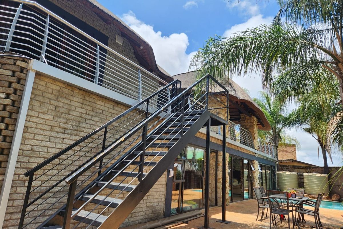 Exterior rear view featuring a thatched roof, brick facade, metal staircase, and a paved patio area overlooking the swimming pool.