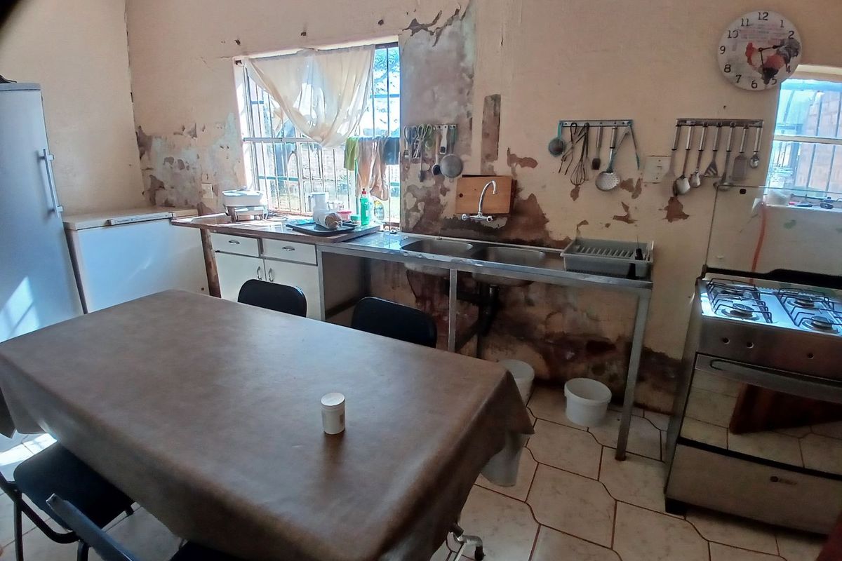 A functional kitchen area featuring a stainless steel sink unit, tiled flooring, and a freestanding stove, requiring significant wall repairs and cosmetic updates.