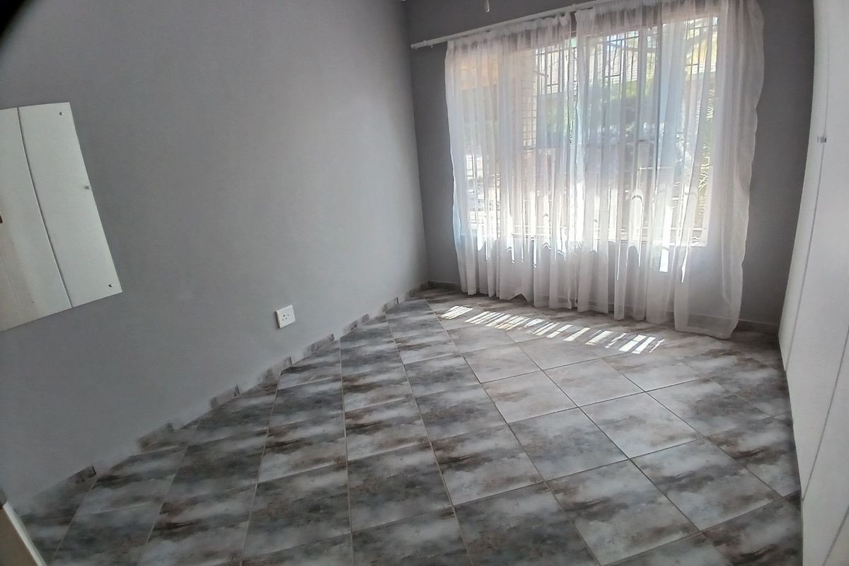 A bright, empty bedroom featuring neutral grey walls, tiled flooring, and large windows with fitted curtain tracks.