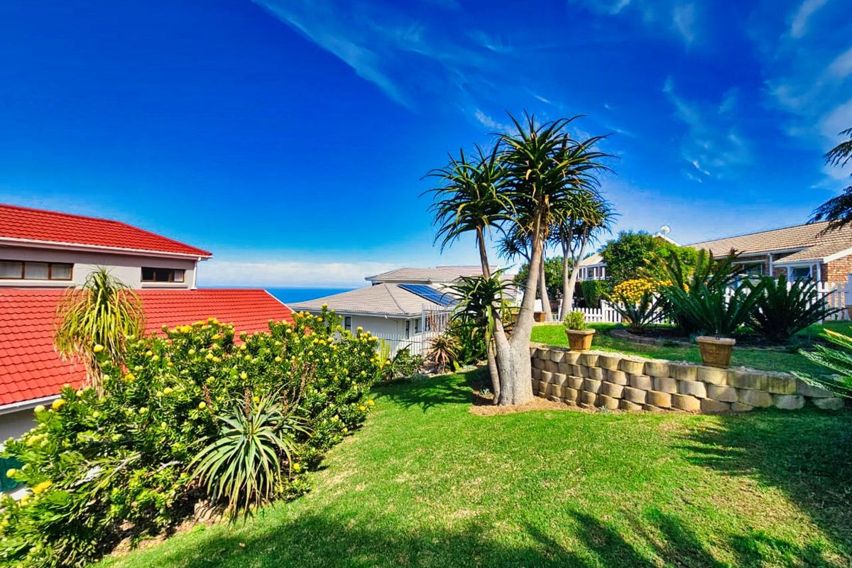 A well-maintained rear garden featuring tiered landscaping, a retaining wall, and mature indigenous plants with a view of the ocean in the distance.