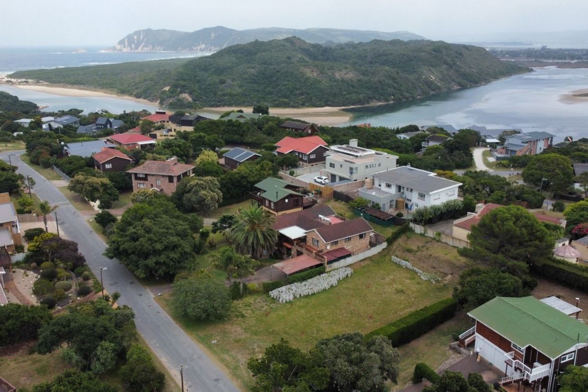 An aerial view of a residential neighborhood featuring a vacant plot of land, surrounded by established homes, lush greenery, and nearby coastal landscape.