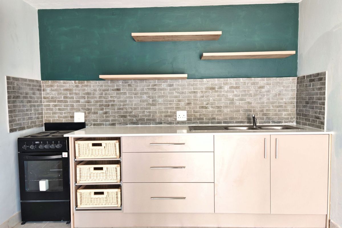 A compact kitchen featuring modern base cabinetry, a stainless steel sink, a built-in oven with hob, and decorative floating shelves against a tiled backsplash.