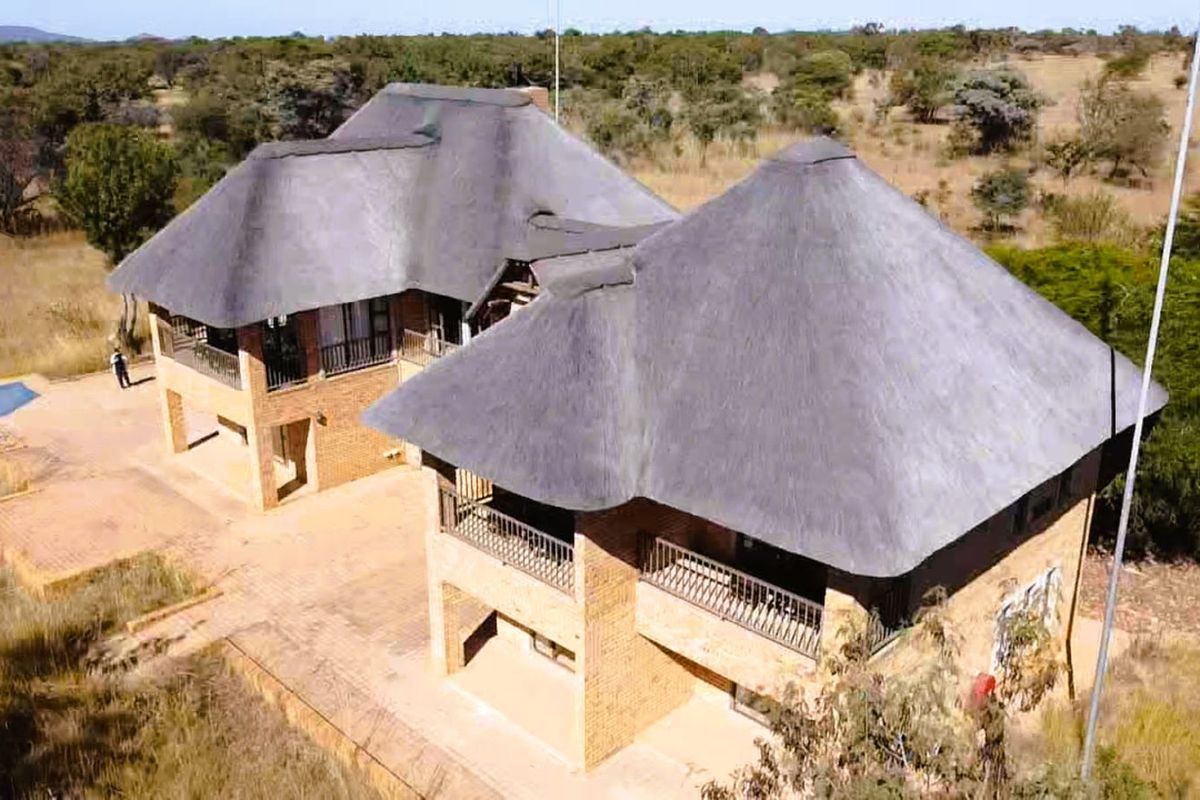 Aerial view of a multi-level brick house featuring traditional thatched roofing, wrap-around balconies, and an adjacent swimming pool in a rural landscape.
