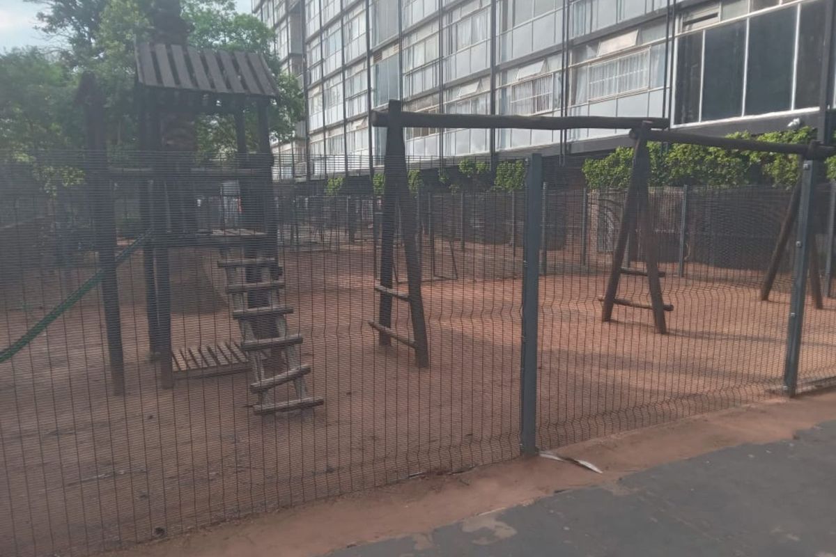 A communal outdoor play area featuring wooden climbing structures and swings, enclosed by security fencing within an apartment complex setting.