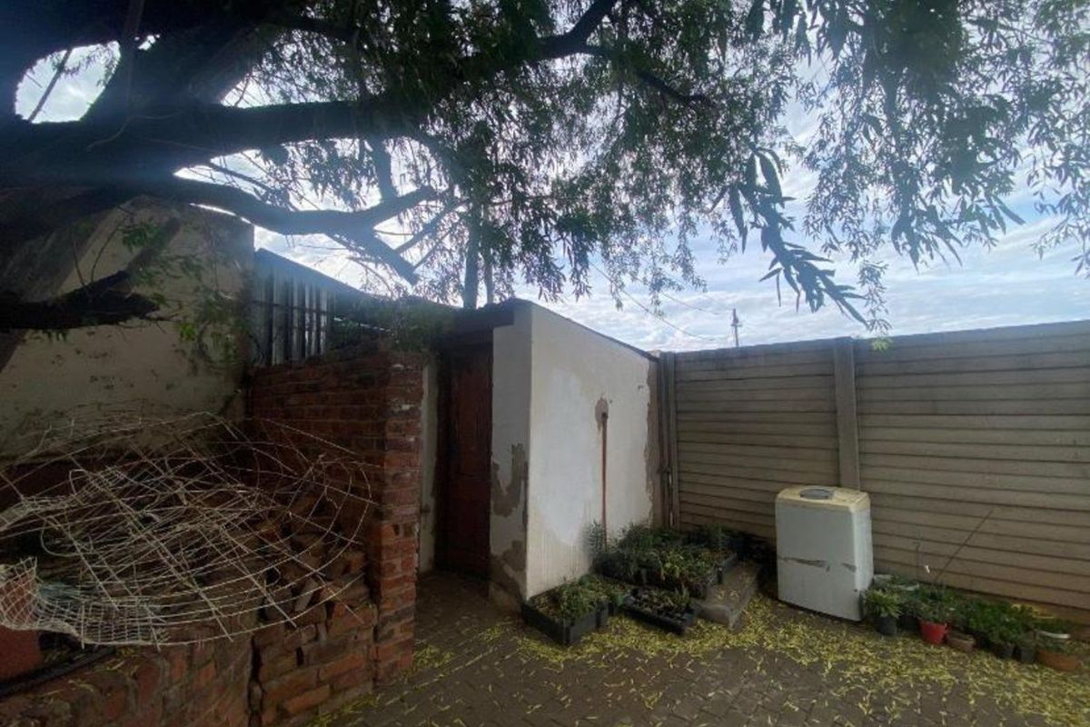 A rear outdoor area featuring a paved courtyard, brick wall structure, and perimeter fencing, showing signs of significant wear and weathering.