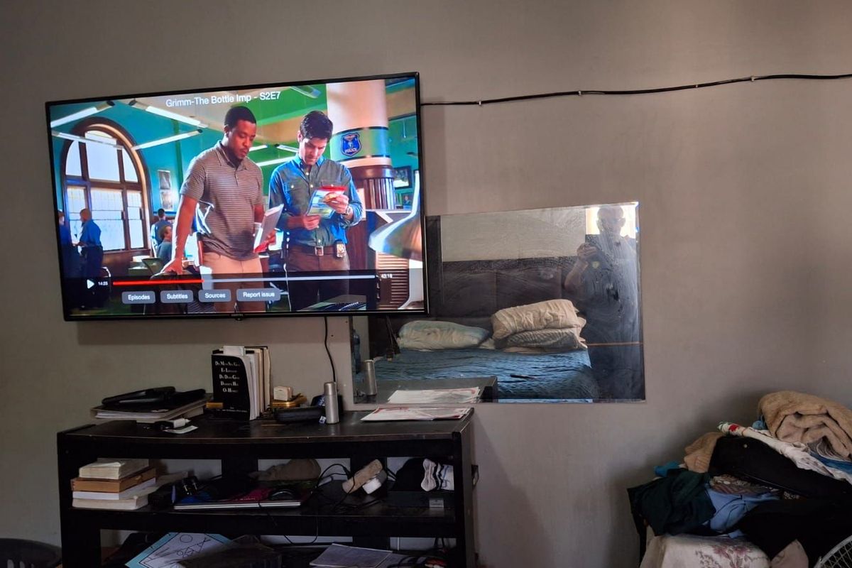 A bedroom interior featuring a wall-mounted television and a mirror, with visible exposed wiring and basic wall finishes.