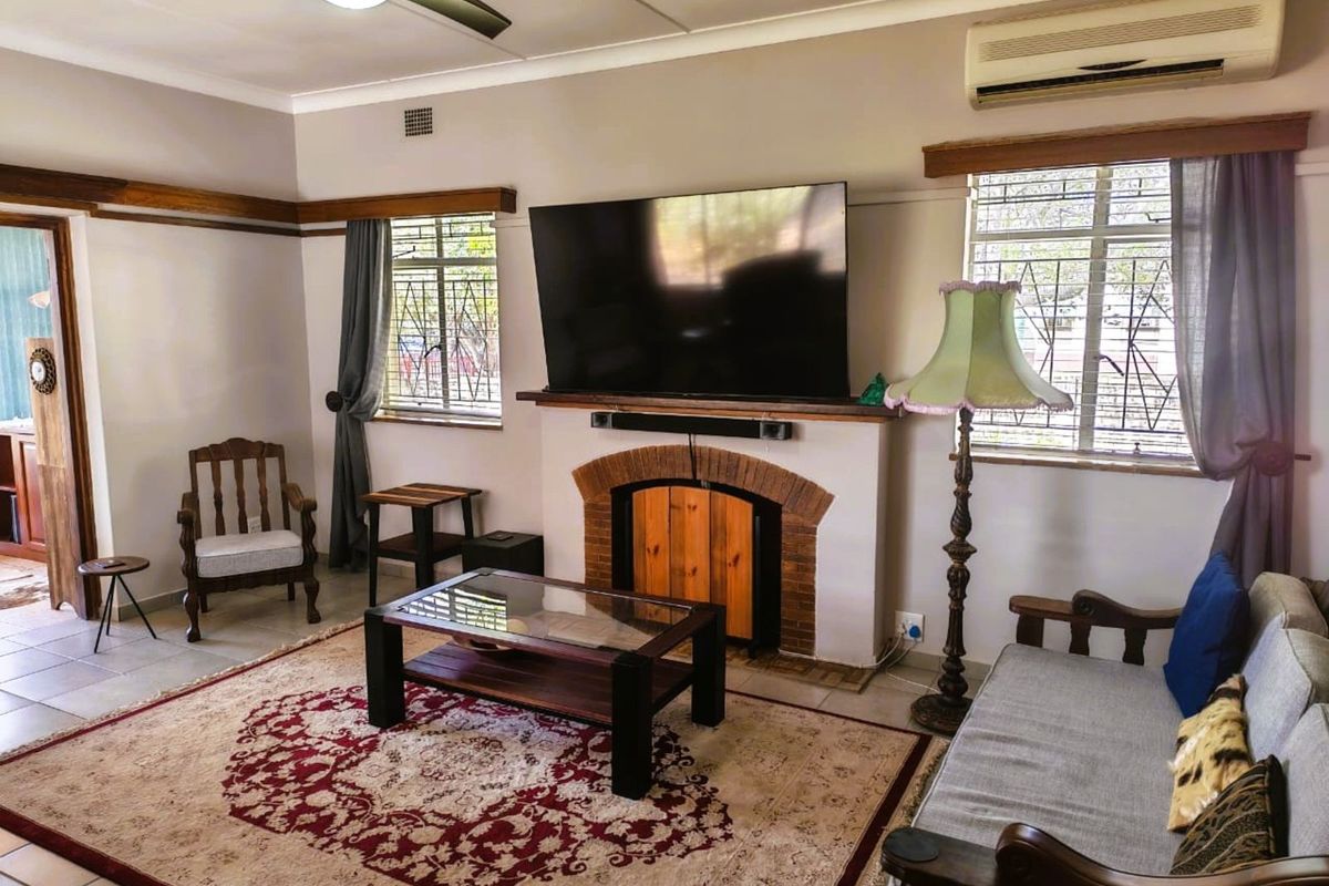 A comfortable living room featuring a central brick fireplace, ceiling fan, and wall-mounted air conditioning unit, with large windows providing natural light.