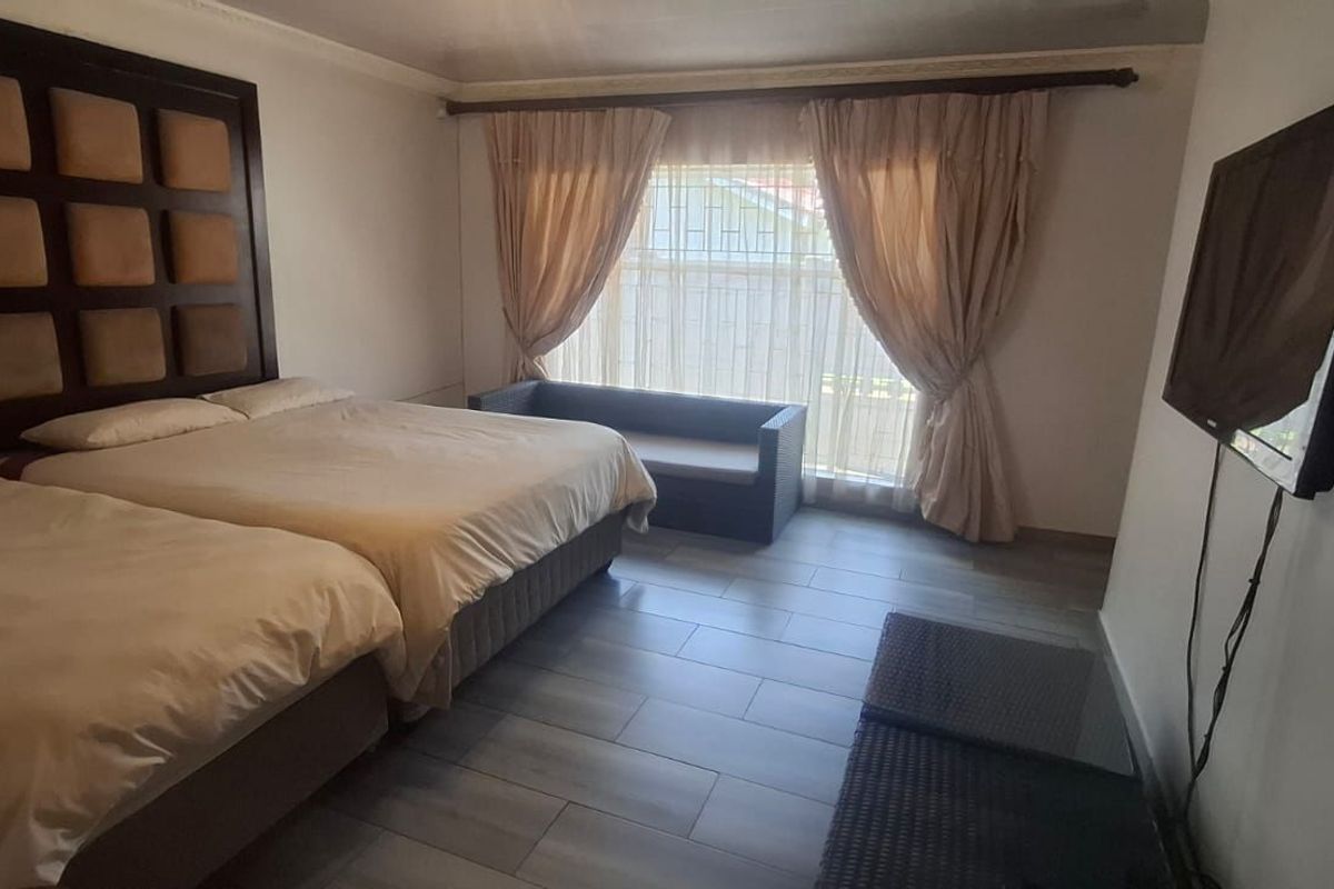 A spacious bedroom featuring tiled flooring, a large window with curtains, and a wall-mounted television.