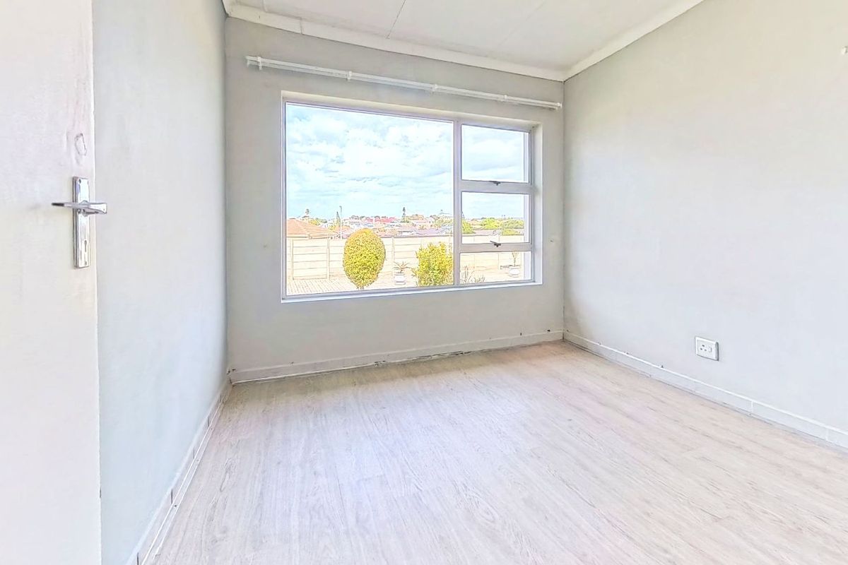 A bright, empty bedroom featuring light-toned laminate flooring, a large window with natural light, and neutral wall finishes.