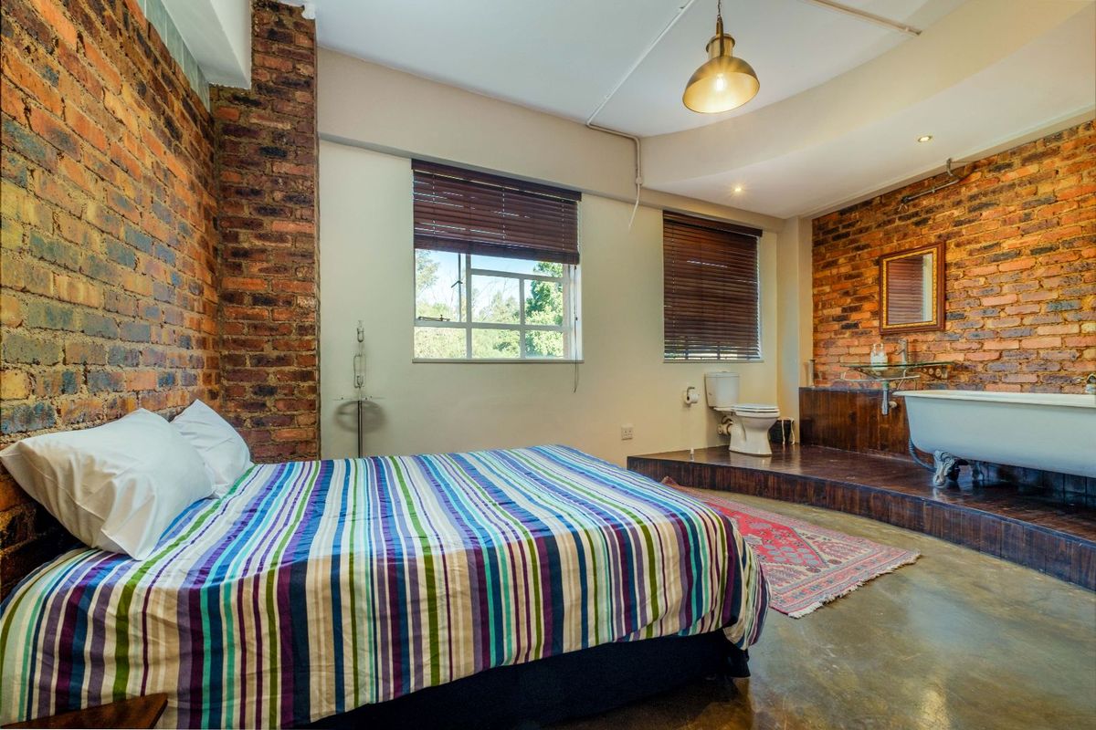 A unique bedroom featuring an open-plan en-suite bathroom with a clawfoot bathtub, exposed brick walls, and polished concrete flooring.