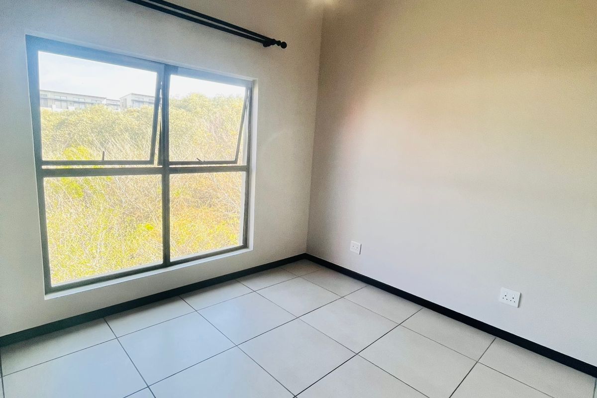 A bright, empty bedroom featuring neutral wall tones, large window with natural light, and clean tiled flooring.