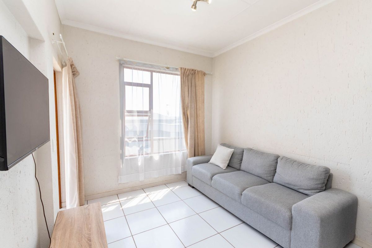 A compact living room featuring white tiled flooring, textured walls, and a large window allowing natural light into the space.