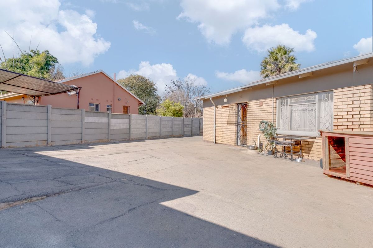 A spacious rear exterior area featuring a concrete driveway, perimeter boundary wall, and brick facade with security gate access.