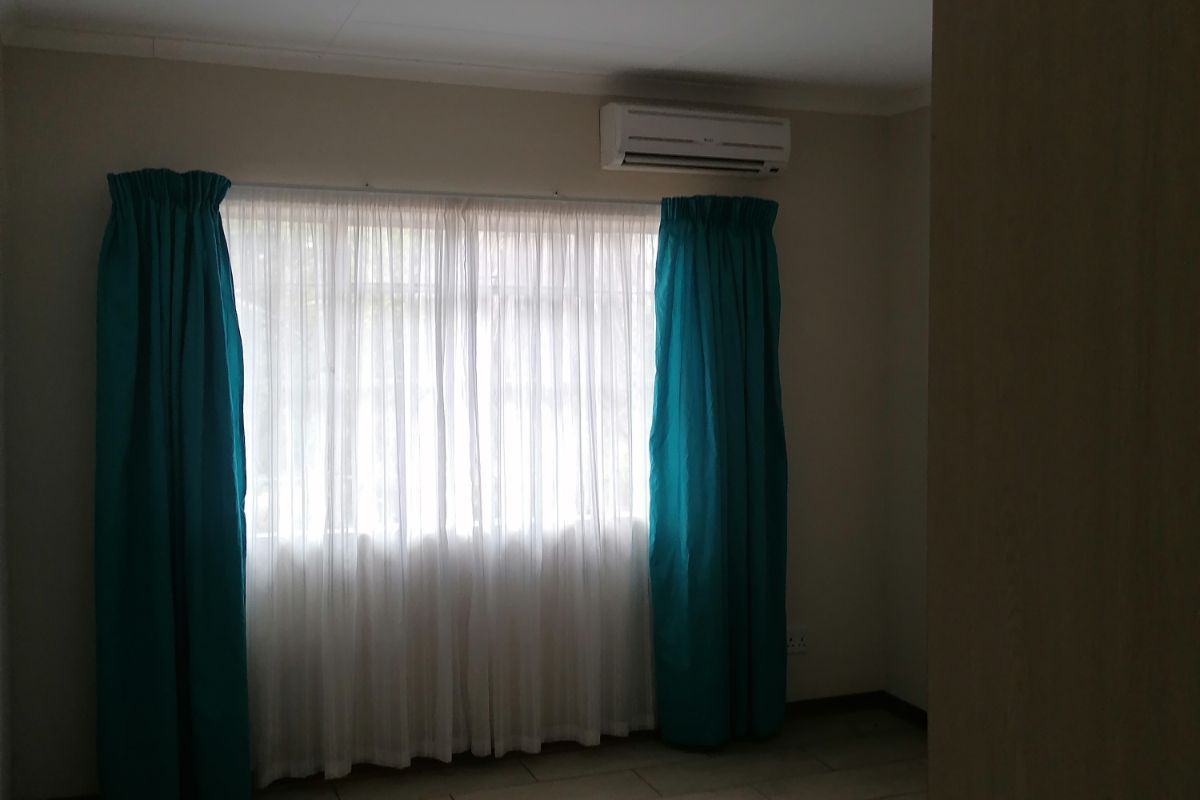 A bright bedroom featuring tiled flooring, a wall-mounted air conditioning unit, and large windows with curtains.
