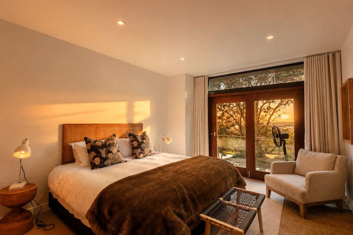 A well-appointed bedroom featuring recessed ceiling lighting, neutral carpeting, and large timber-framed glass doors opening to a balcony with scenic sunset views.