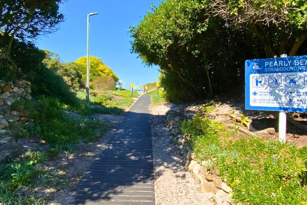 A wooden boardwalk path leading through a landscaped coastal area towards the beach, featuring stone retaining walls and resort signage.