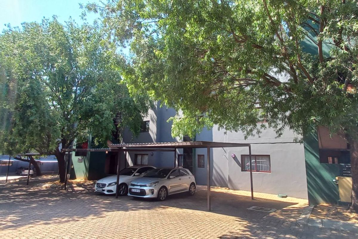 Exterior view of the apartment complex featuring paved parking areas, covered carports, and mature trees surrounding the building facade.