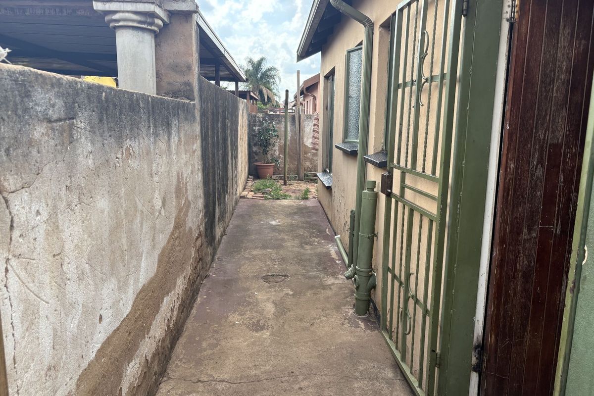A narrow side passage featuring concrete paving, a security gate, and a weathered boundary wall requiring maintenance.