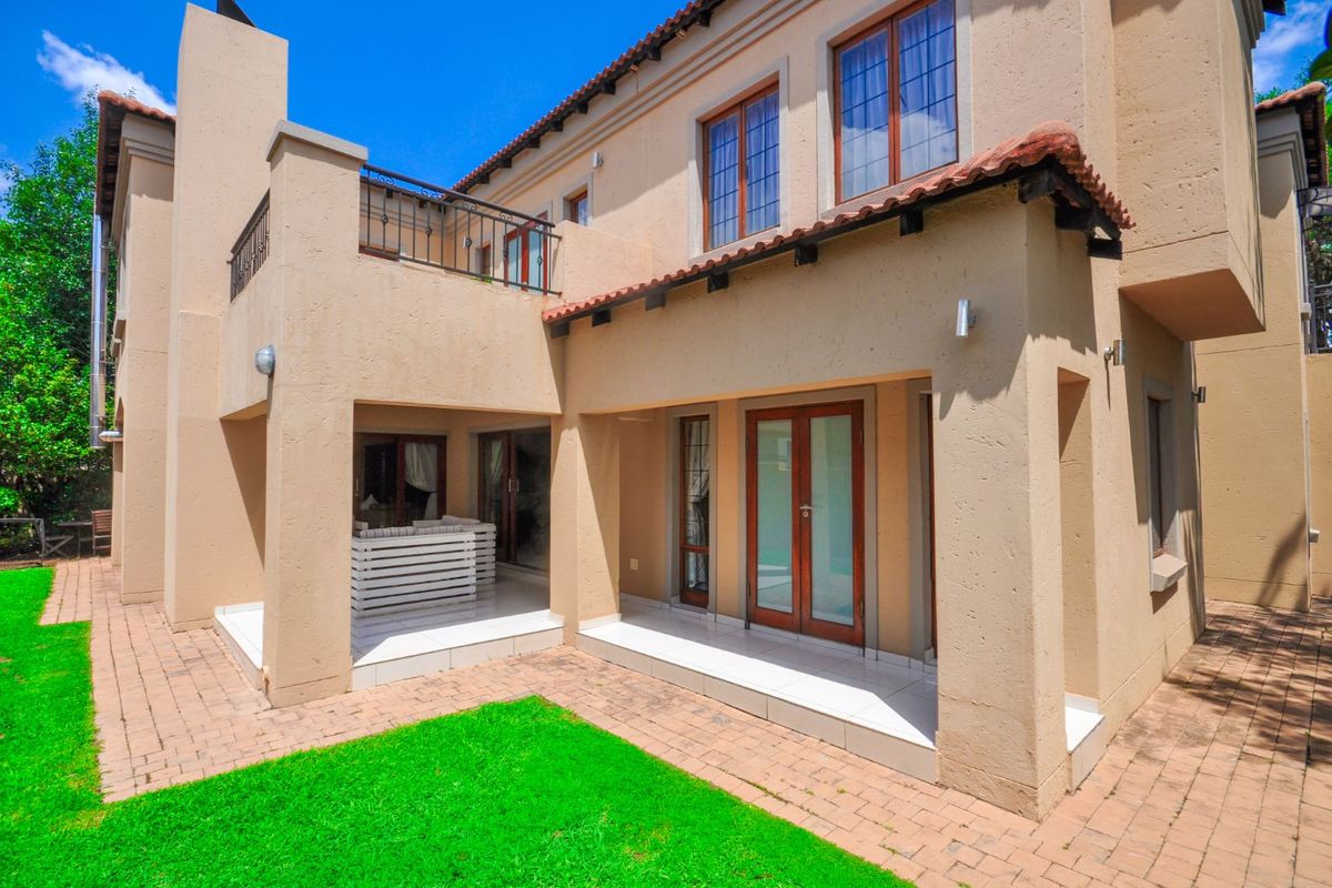 Rear view of a two-story house featuring a covered patio, tiled flooring, balcony with wrought iron railing, and a paved garden area.