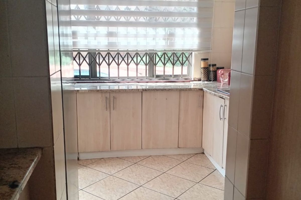 A functional kitchen featuring light wood-finish cabinetry, granite countertops, tiled flooring, and a window with security bars and a roller blind.