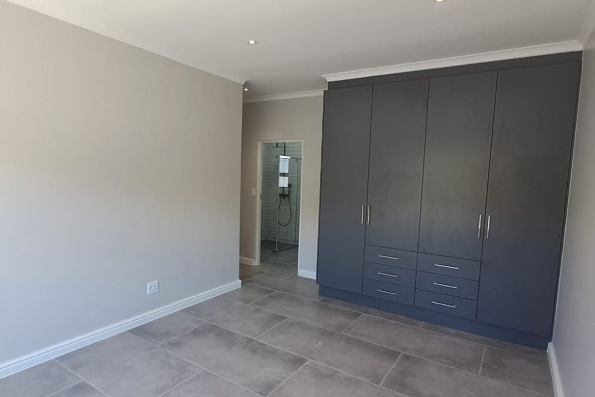 A modern bedroom featuring large format floor tiles, built-in dark grey cabinetry, and an adjoining en-suite bathroom with a glass-enclosed shower.