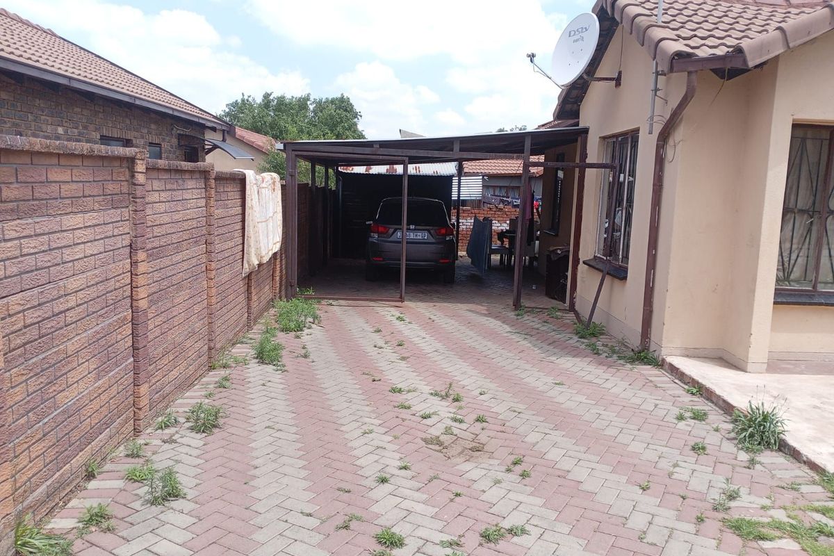 Side exterior view showing a paved driveway, a metal carport structure, and a brick boundary wall.