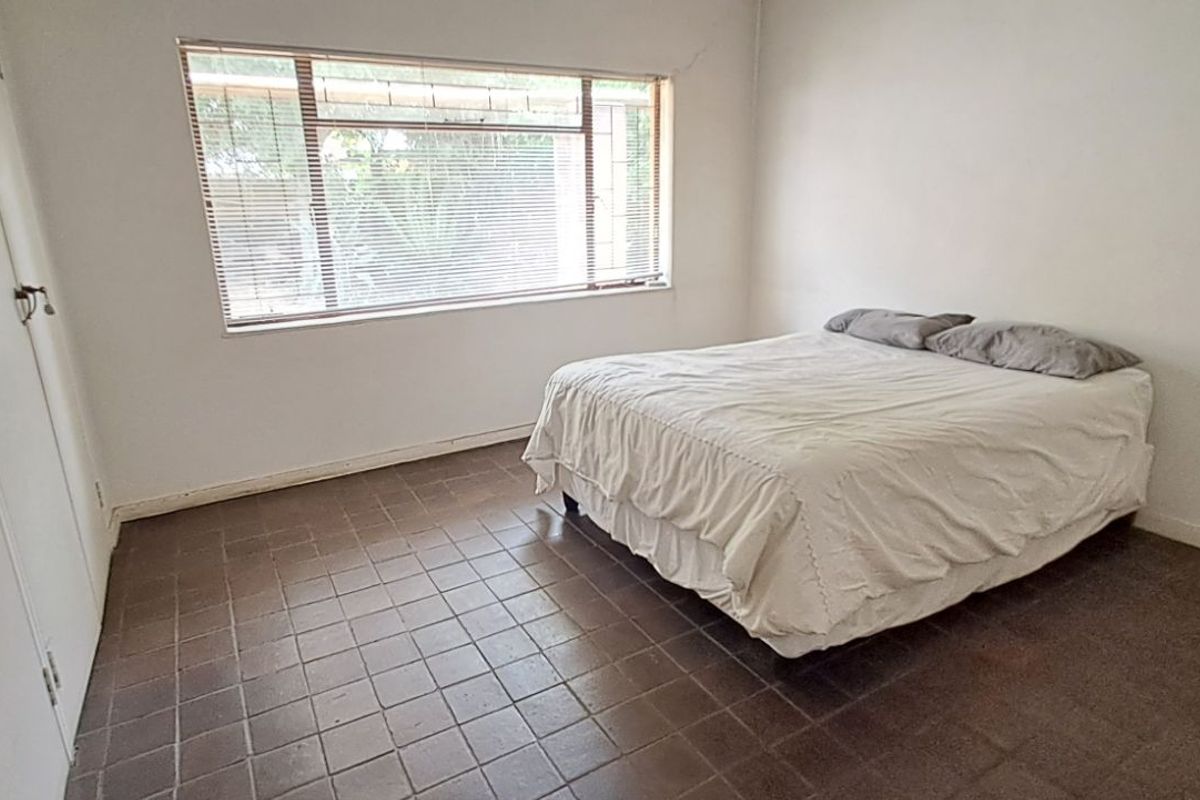 A simple bedroom featuring tiled flooring, built-in cupboards, and a large window with horizontal blinds.
