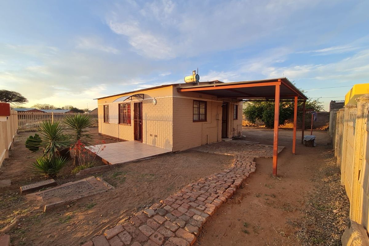Exterior view of a single-story house featuring a covered carport, tiled patio area, and a brick-paved pathway leading to the entrance.