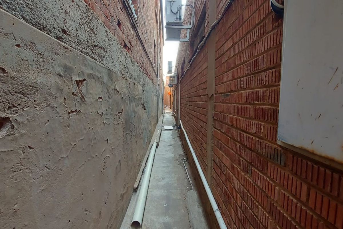 A narrow exterior alleyway between two brick buildings, featuring exposed piping, weathered masonry, and wall-mounted air conditioning units.