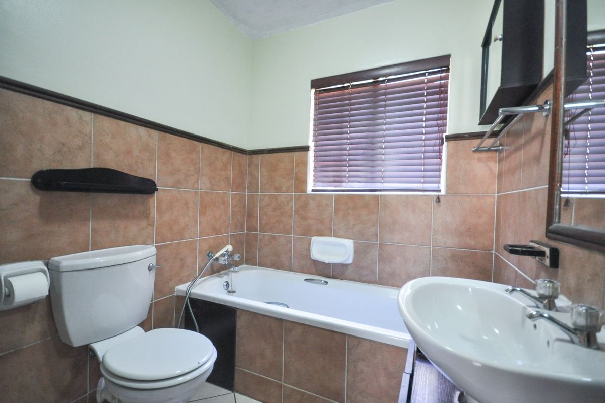 A functional bathroom featuring a built-in bathtub, pedestal sink, and toilet, finished with neutral wall tiles and a window with fitted blinds.