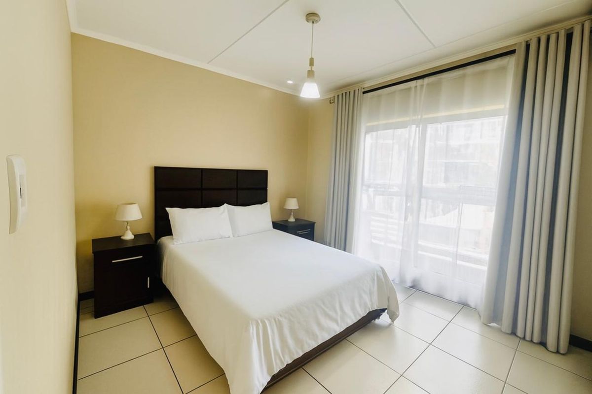 A well-lit bedroom featuring tiled flooring, a large window with curtain tracks, and a central pendant light fixture.