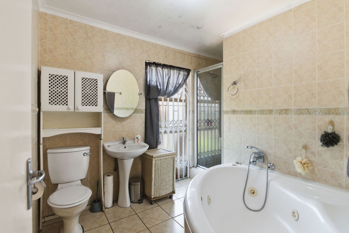 A functional bathroom featuring a corner jetted bathtub, pedestal basin, toilet, and full wall tiling with a security gate at the window.