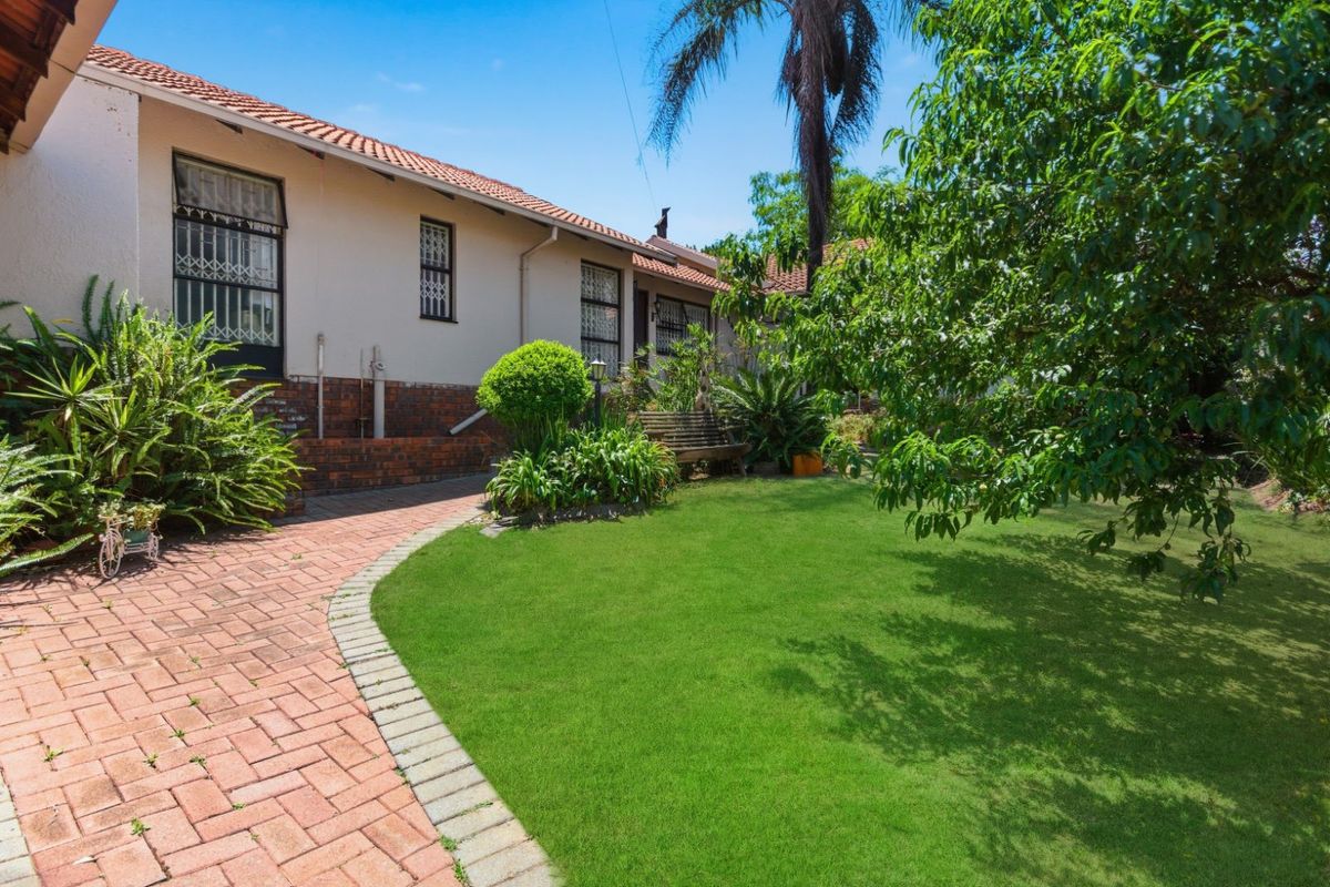 A view of the rear garden featuring a manicured lawn, brick paved pathway, and established trees adjacent to the house exterior.