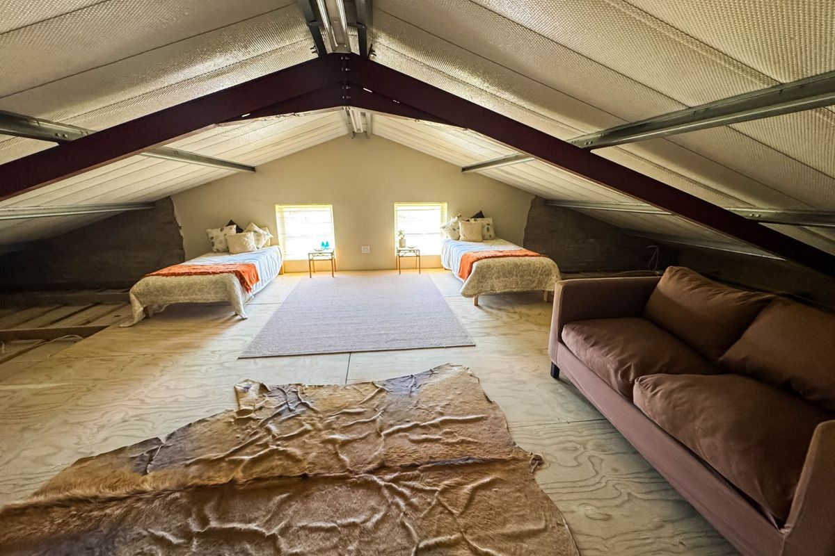 Spacious attic-style bedroom featuring exposed steel roof trusses, plywood flooring, and dual windows, offering a rustic and versatile living space.